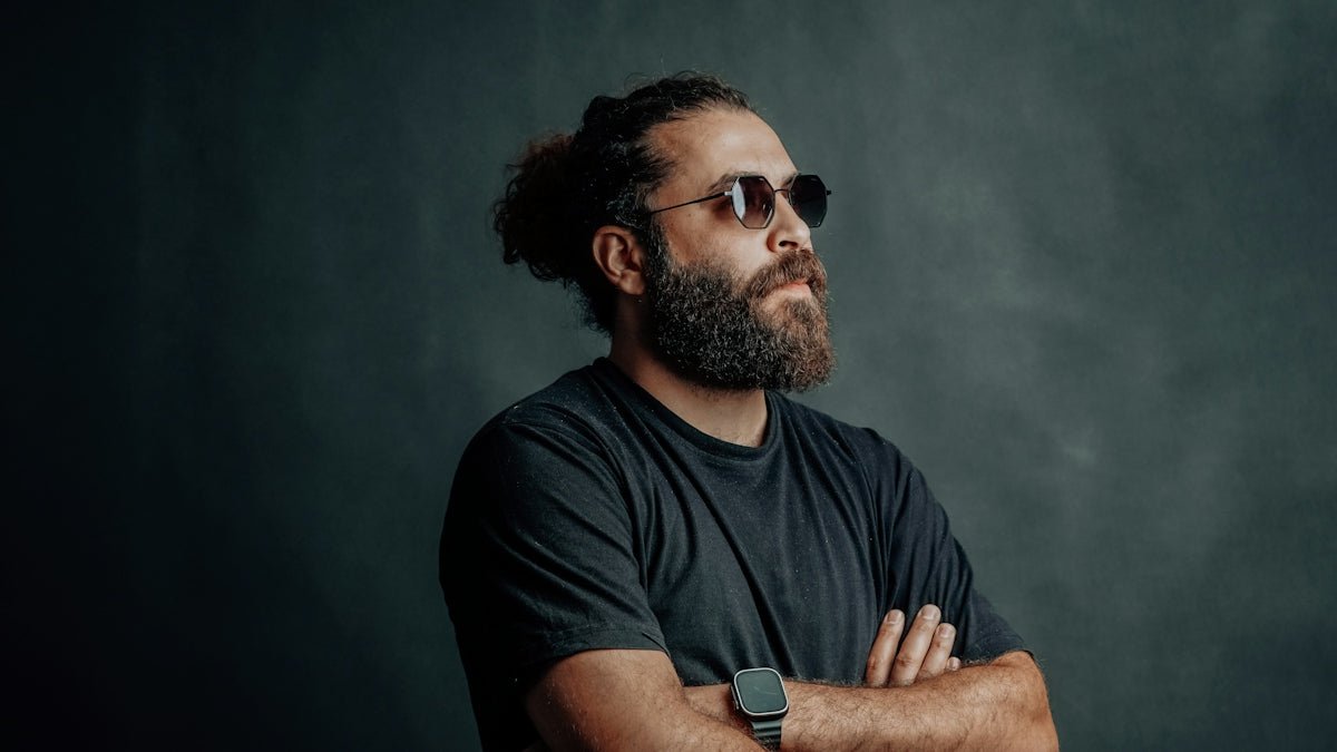 A bearded man wears sunglasses and looks away.