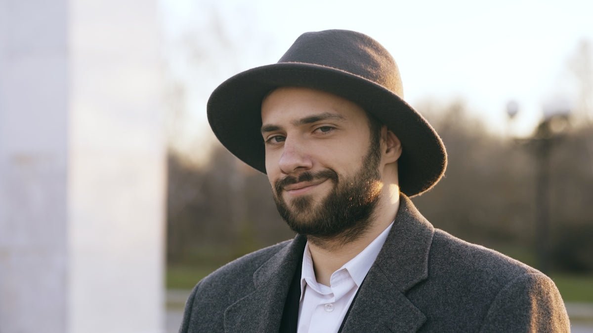 Man wearing a hat and coat smiles outdoors.