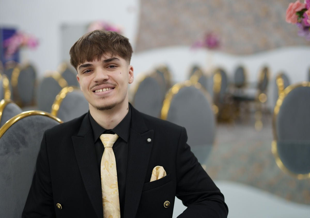 Young man in a black suit and gold tie smiles.