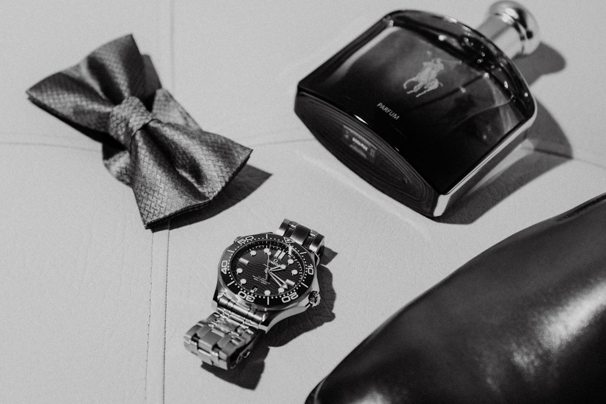 A watch, bowtie, cologne, and shoe arranged neatly.