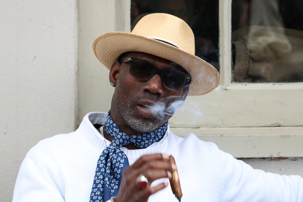 Man wearing a hat and sunglasses smoking a cigar.
